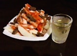 A white plate filled with pieces of jicama and mango, sprinkled with chili powder and tamarind candies on top. Beside the plate, there is a clear glass filled with a chilled beverage that appears to contain ice cubes.