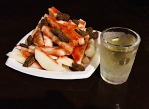 A white plate filled with pieces of jicama and mango, sprinkled with chili powder and tamarind candies on top. Beside the plate, there is a clear glass filled with a chilled beverage that appears to contain ice cubes.