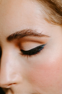 A close-up of a person's face focusing on the eye area, showcasing a perfectly shaped eyebrow and eye makeup with sleek black eyeliner and neutral eyeshadow.