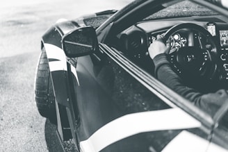 A monochrome photograph of a person driving a sports car. The image captures the driver's hand on the steering wheel and the sleek design of the vehicle's interior. The exterior showcases stylish bodywork with bold lines, and the tires add to the performance-oriented appearance.