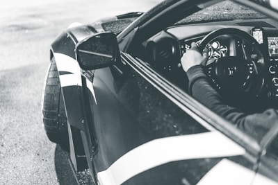 A monochrome photograph of a person driving a sports car. The image captures the driver's hand on the steering wheel and the sleek design of the vehicle's interior. The exterior showcases stylish bodywork with bold lines, and the tires add to the performance-oriented appearance.