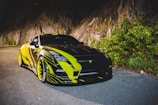 A sleek sports car with a black and yellow design is parked on a road adjacent to a rocky cliffside. The car features bold lines and patterns, exuding a dynamic appearance. The surrounding area includes greenery and a rugged rock face which adds to the adventurous feel.