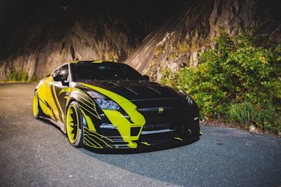 A sleek sports car with a black and yellow design is parked on a road adjacent to a rocky cliffside. The car features bold lines and patterns, exuding a dynamic appearance. The surrounding area includes greenery and a rugged rock face which adds to the adventurous feel.