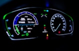 A digital car dashboard displaying various indicators and metrics including speed, power, remaining kilometers, fuel consumption in liters per 100 kilometers, and temperature. The dashboard is illuminated with neon-like blue and green lights, showing additional symbols and alerts related to driving mode and car status.
