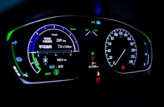 A digital car dashboard displaying various indicators and metrics including speed, power, remaining kilometers, fuel consumption in liters per 100 kilometers, and temperature. The dashboard is illuminated with neon-like blue and green lights, showing additional symbols and alerts related to driving mode and car status.