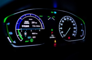 A digital car dashboard displaying various indicators and metrics including speed, power, remaining kilometers, fuel consumption in liters per 100 kilometers, and temperature. The dashboard is illuminated with neon-like blue and green lights, showing additional symbols and alerts related to driving mode and car status.