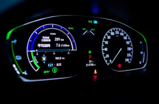 A digital car dashboard displaying various indicators and metrics including speed, power, remaining kilometers, fuel consumption in liters per 100 kilometers, and temperature. The dashboard is illuminated with neon-like blue and green lights, showing additional symbols and alerts related to driving mode and car status.