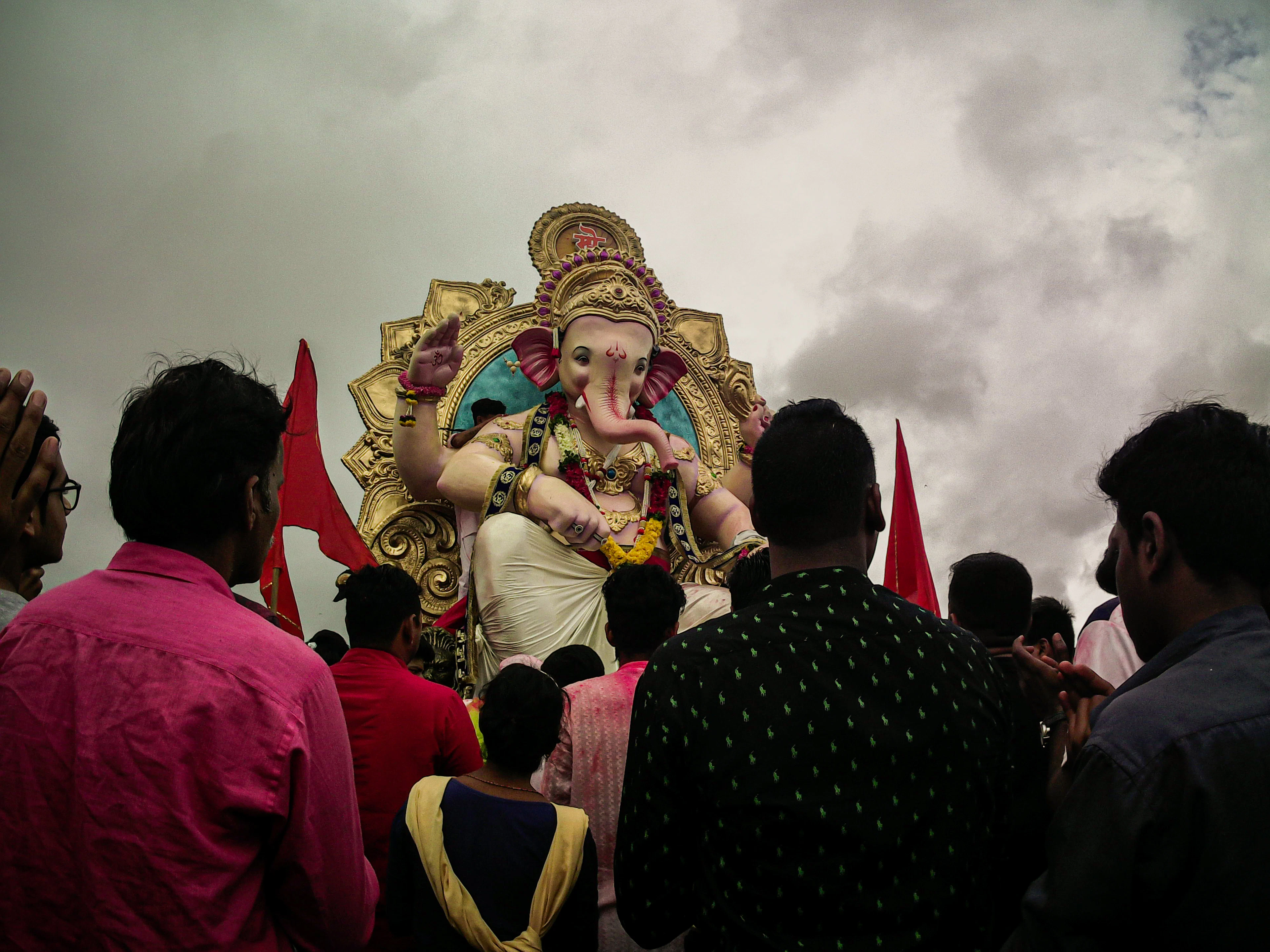 A large Ganesh idol is carried through a crowd of devotees under a stormy grey sky during a festival procession.