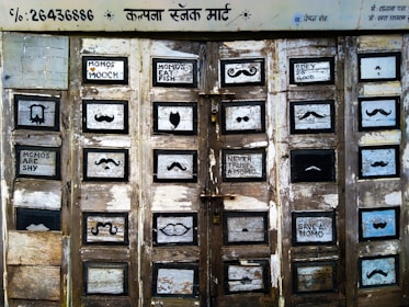 A series of old, weathered wooden doors with multiple small, square panels, each displaying various mustache designs and whimsical text messages. The paint is peeling, giving an antique appearance. Hindi writing is visible at the top, and each panel features either a painted mustache style or humorous phrases like 'Momos Mooch' and 'Momos Are Shy'.