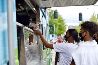 A transaction at a food truck as a person hands an item to a customer through the service window. Two people wearing face masks stand at the truck, one of them reaching out to receive their order. The setting is outdoors with blurred people and trees in the background, suggesting a lively and public atmosphere.