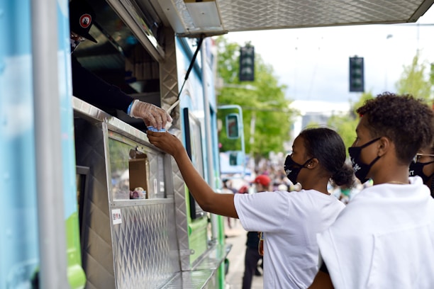 A transaction at a food truck as a person hands an item to a customer through the service window. Two people wearing face masks stand at the truck, one of them reaching out to receive their order. The setting is outdoors with blurred people and trees in the background, suggesting a lively and public atmosphere.