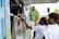 A transaction at a food truck as a person hands an item to a customer through the service window. Two people wearing face masks stand at the truck, one of them reaching out to receive their order. The setting is outdoors with blurred people and trees in the background, suggesting a lively and public atmosphere.