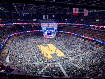 A packed basketball arena with a center court surrounded by thousands of spectators in the stands. The large jumbotron displays game information, and flags hang from the ceiling. The arena is well-lit, with banners running along the upper edges promoting various messages.