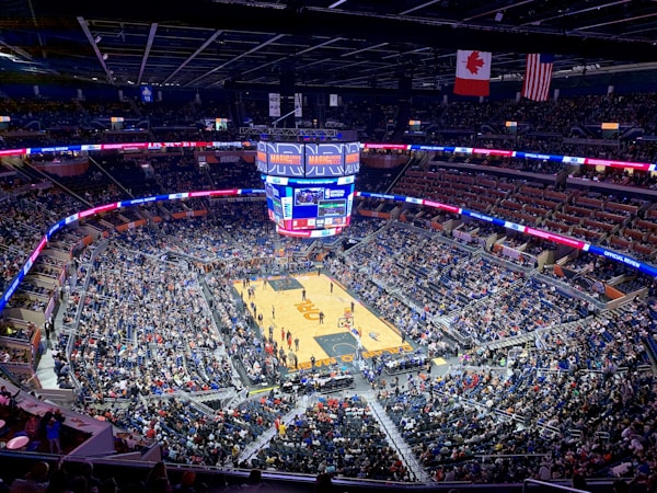 A packed basketball arena with a center court surrounded by thousands of spectators in the stands. The large jumbotron displays game information, and flags hang from the ceiling. The arena is well-lit, with banners running along the upper edges promoting various messages.