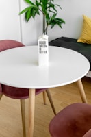 A white round table with wooden legs is placed in a room with a carton labeled 'Boxed Water Is Better' positioned at the center. Two pink velvet chairs are partially visible around the table. A green houseplant is placed in the background near a white wall, and a yellow cushion is on a dark sofa.