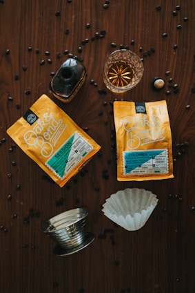 Two packets of Oak & Bond Coffee Co. coffee are placed on a wooden surface with scattered coffee beans. There is a small glass of amber liquid, possibly whiskey or liqueur, next to a dark bottle. A metallic coffee filter holder and a paper filter are also present.