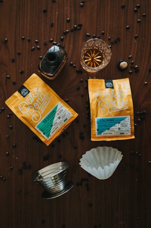 Two packets of Oak & Bond Coffee Co. coffee are placed on a wooden surface with scattered coffee beans. There is a small glass of amber liquid, possibly whiskey or liqueur, next to a dark bottle. A metallic coffee filter holder and a paper filter are also present.