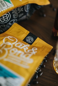 A close-up of a yellow coffee bag with the logo 'Oak & Bond Coffee'. The focus is on the branding, with coffee beans scattered around on a wooden table, creating a rustic and inviting atmosphere.