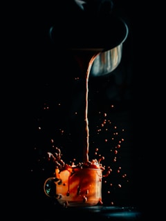 A stream of liquid is being poured from a pot into a glass cup, causing a dynamic splash with droplets suspended in mid-air. The scene is low-lit, highlighting the liquid's rich color against a dark background.