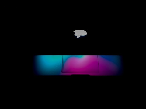 A laptop with an illuminated apple logo against a dark background. The keyboard area emits a vibrant, colorful glow featuring shades of blue, pink, and purple.
