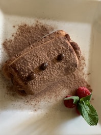 A rectangular dessert coated with cocoa powder is garnished with coffee beans on top. The dessert is complemented by fresh raspberries and a sprig of mint on the side, all arranged on a white plate.