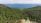 A dense, lush green forest covers the hillside, descending towards a small, isolated beach with turquoise waters. A series of small red-roofed cabins is nestled at the edge of the forest near the coastline. In the distance, a calm sea stretches out to the horizon, with a small island visible far off.