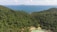 A dense, lush green forest covers the hillside, descending towards a small, isolated beach with turquoise waters. A series of small red-roofed cabins is nestled at the edge of the forest near the coastline. In the distance, a calm sea stretches out to the horizon, with a small island visible far off.