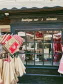 A quaint shop display featuring textiles such as oven mitts and tablecloths with red and beige designs. The storefront is adorned with a window reflecting various objects and part of the street. The signage above the window reads 'Boutique sur 3 niveaux' in elegant script.