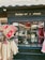 A quaint shop display featuring textiles such as oven mitts and tablecloths with red and beige designs. The storefront is adorned with a window reflecting various objects and part of the street. The signage above the window reads 'Boutique sur 3 niveaux' in elegant script.