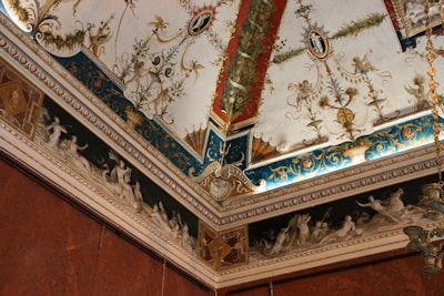 An intricately decorated ceiling corner with ornate moldings and paintings. The design includes classical motifs, floral patterns, and detailed frescoes depicting figures. Rich colors and gilded elements enhance the luxurious and artistic appeal.