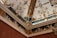 An intricately decorated ceiling corner with ornate moldings and paintings. The design includes classical motifs, floral patterns, and detailed frescoes depicting figures. Rich colors and gilded elements enhance the luxurious and artistic appeal.