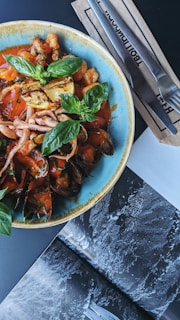 A colorful seafood dish is presented in a blue rimmed bowl, featuring mussels, squid, shrimp, and baby octopus in a red tomato sauce with fresh basil leaves and grilled lemon garnishes. Next to the bowl, a set of cutlery rests on paper napkins, and part of a black and white open magazine is visible on the table.