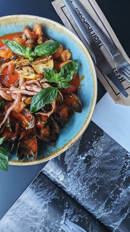 A colorful seafood dish is presented in a blue rimmed bowl, featuring mussels, squid, shrimp, and baby octopus in a red tomato sauce with fresh basil leaves and grilled lemon garnishes. Next to the bowl, a set of cutlery rests on paper napkins, and part of a black and white open magazine is visible on the table.