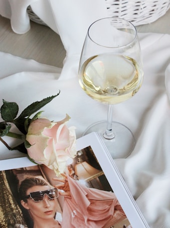 A white wine glass is elegantly placed on a smooth, white fabric surface. Nearby, a delicate pink rose adds a touch of softness next to an open magazine featuring a stylishly dressed woman wearing sunglasses. The setting is serene and refined, with a wicker basket subtly visible in the background.