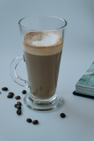 A tall glass filled with a frothy latte stands on a light surface, surrounded by scattered coffee beans. To the right, a closed book with a patterned cover edges into the frame. The overall setting suggests a calm and cozy atmosphere, likely intended for leisurely coffee enjoyment.