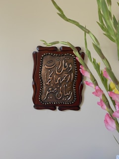 A decorative wooden plaque with Arabic calligraphy is mounted on a beige wall. The text appears to be embossed with metallic elements, framed in an ornate design. On the right side of the image, vibrant pink gladiolus flowers with green leaves are partially visible.