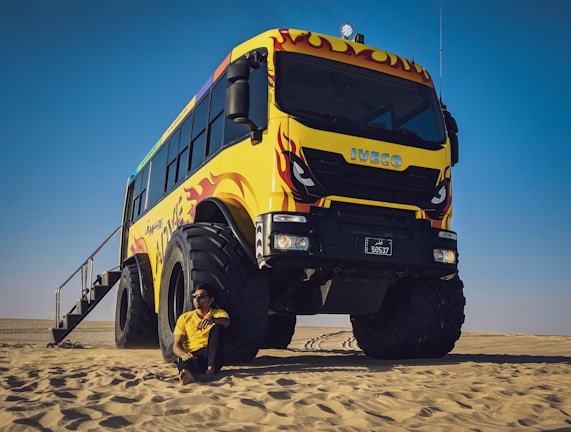 A large, colorful off-road bus with vibrant yellow and red flame designs is parked in a desert setting. A man in a yellow shirt is sitting on the sand next to the bus, under a clear blue sky.
