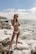 A person wearing a white bikini stands on rocky shore with waves crashing around them. The ocean stretches out to the horizon under a partly cloudy blue sky. Sunglasses complement the casual and relaxed beach vibe.