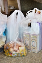Several plastic bags filled with groceries are placed on a speckled surface. The bags contain various items, including two cartons featuring a person's portrait, a bag filled with eggs packaged in a transparent plastic bag, and other items obscured by the bags. A pair of slippers is visible in the background.