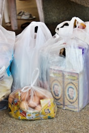 Several plastic bags filled with groceries are placed on a speckled surface. The bags contain various items, including two cartons featuring a person's portrait, a bag filled with eggs packaged in a transparent plastic bag, and other items obscured by the bags. A pair of slippers is visible in the background.
