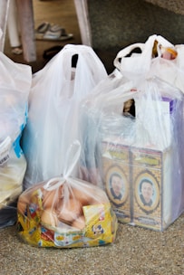 Several plastic bags filled with groceries are placed on a speckled surface. The bags contain various items, including two cartons featuring a person's portrait, a bag filled with eggs packaged in a transparent plastic bag, and other items obscured by the bags. A pair of slippers is visible in the background.