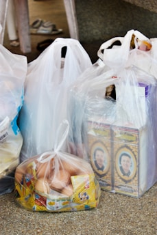 Several plastic bags filled with groceries are placed on a speckled surface. The bags contain various items, including two cartons featuring a person's portrait, a bag filled with eggs packaged in a transparent plastic bag, and other items obscured by the bags. A pair of slippers is visible in the background.