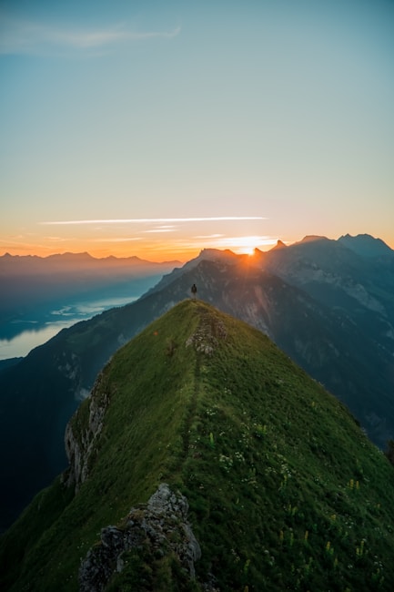 A solitary figure stands at the peak of a lush green mountain, observing the sunrise. The landscape features rolling hills and distant mountain ranges under a clear sky. A serene body of water is visible on the left with sunlight casting a golden hue over the scene.