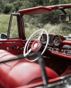 The image features the interior of a classic convertible car with a prominent red and white color scheme. The dashboard is equipped with analog dials and minimalistic controls, evoking a vintage feel. The surroundings outside the car show lush greenery, enhancing the serene and timeless atmosphere.