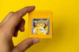 A hand holding a yellow Game Boy cartridge with a vibrant Pokémon label featuring Pikachu on a solid yellow background.