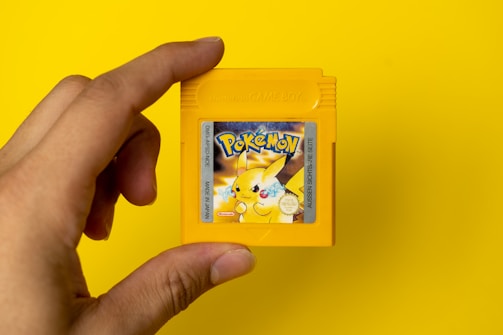 A hand holding a yellow Game Boy cartridge with a vibrant Pokémon label featuring Pikachu on a solid yellow background.