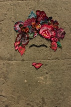 A minimalist artwork on a textured surface resembling a face using vibrant embroidered floral patterns for the hair, a black unibrow, and lips made of pink and yellow thread.