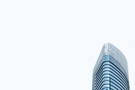 A modern high-rise office building with a sleek glass exterior. The architectural design features curved lines and a minimalist aesthetic. The building stands prominently against a pale, light gray sky, emphasizing its height and contemporary style.