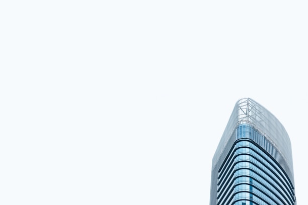A modern high-rise office building with a sleek glass exterior. The architectural design features curved lines and a minimalist aesthetic. The building stands prominently against a pale, light gray sky, emphasizing its height and contemporary style.