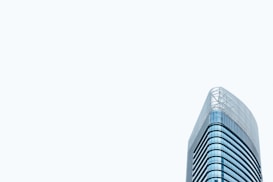 A modern high-rise office building with a sleek glass exterior. The architectural design features curved lines and a minimalist aesthetic. The building stands prominently against a pale, light gray sky, emphasizing its height and contemporary style.